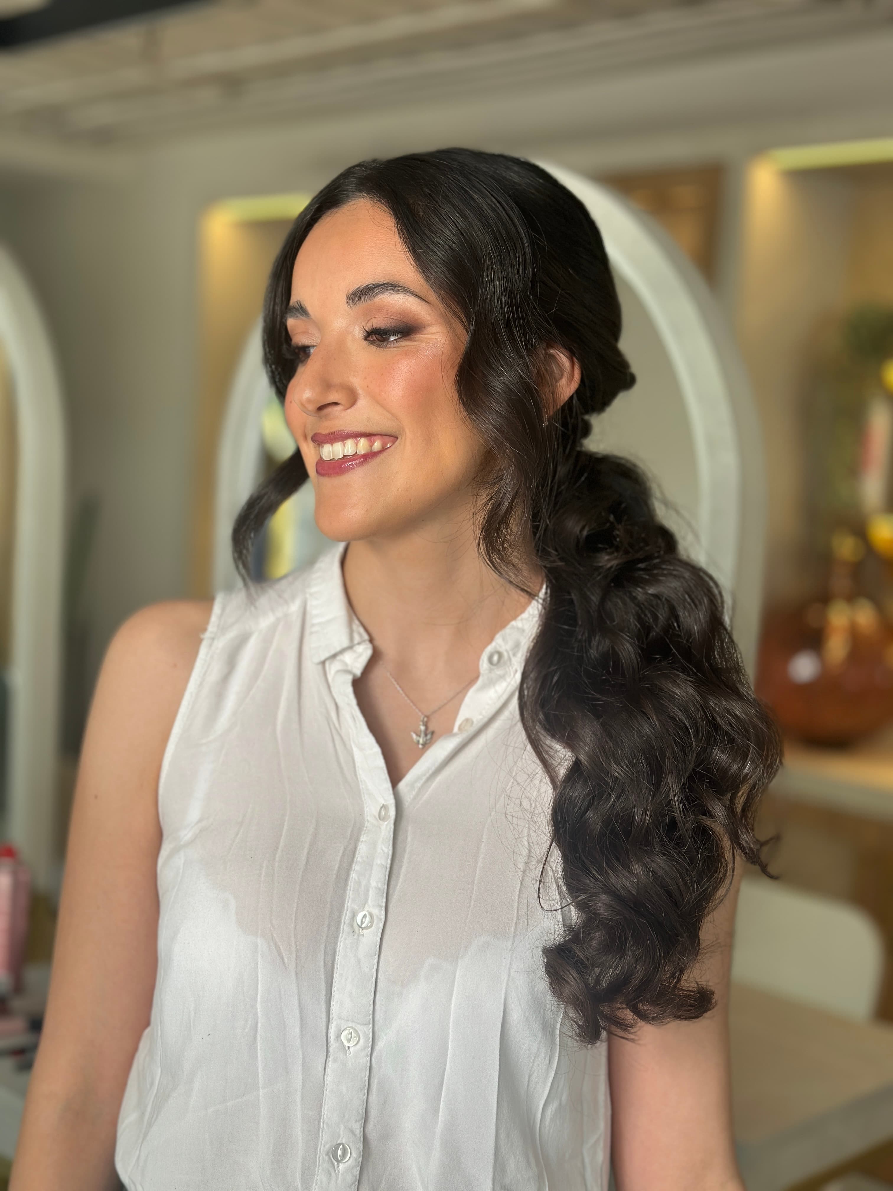 Invitada con cola ondulada, blusa blanca y maquillaje de evento en el estudio